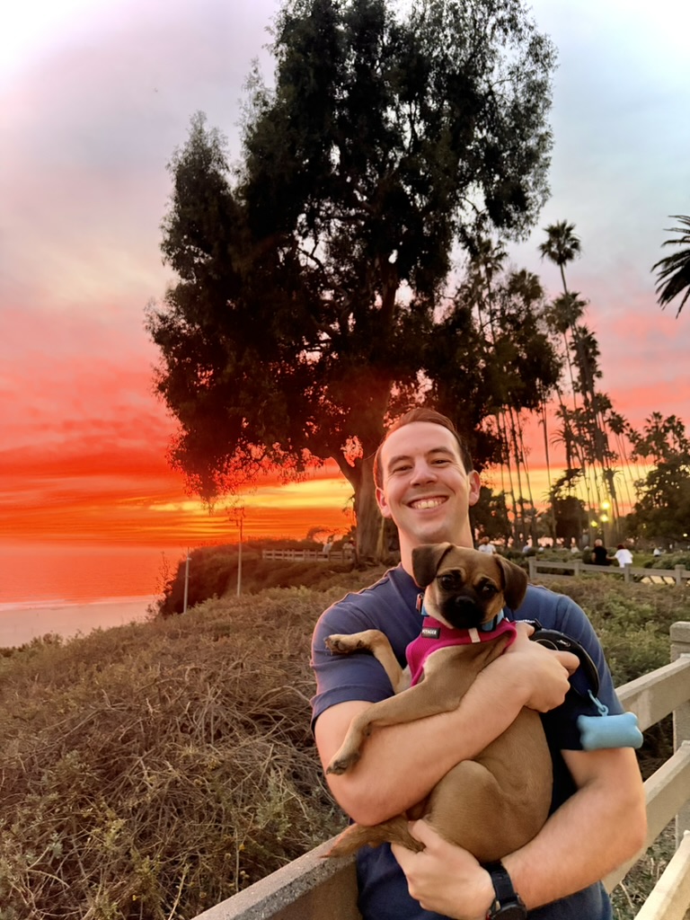 Zachary smiling and holding his small brown dog outdoors at a bright orange sunset by the ocean